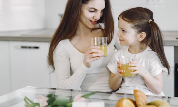 Happy mother and daughter in casual clothes drinking juice and looking at each other while having breakfast together in contemporary kitchen